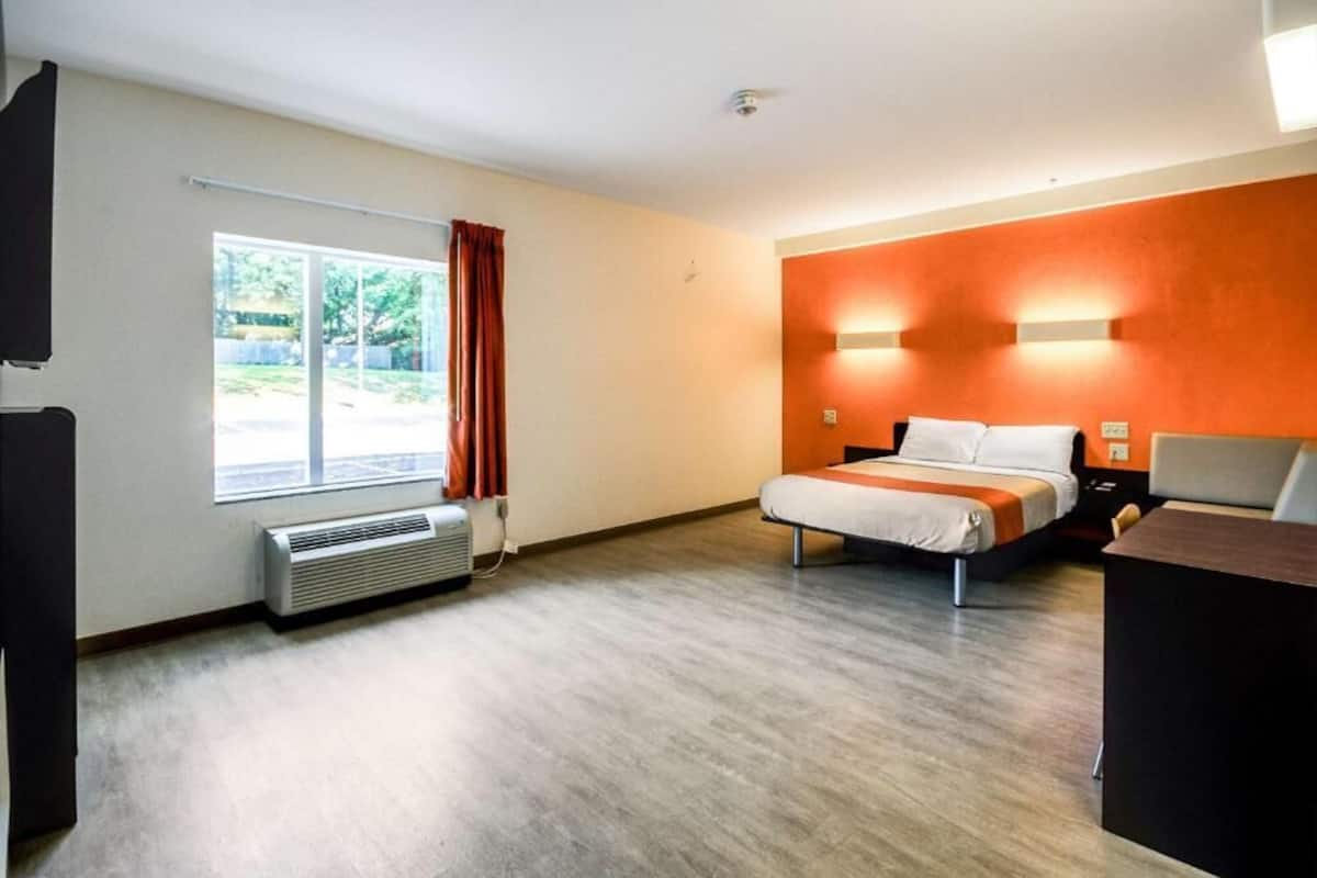 A hotel room with a bed, a desk, a heater, a window with curtains, and a view of the outdoors.