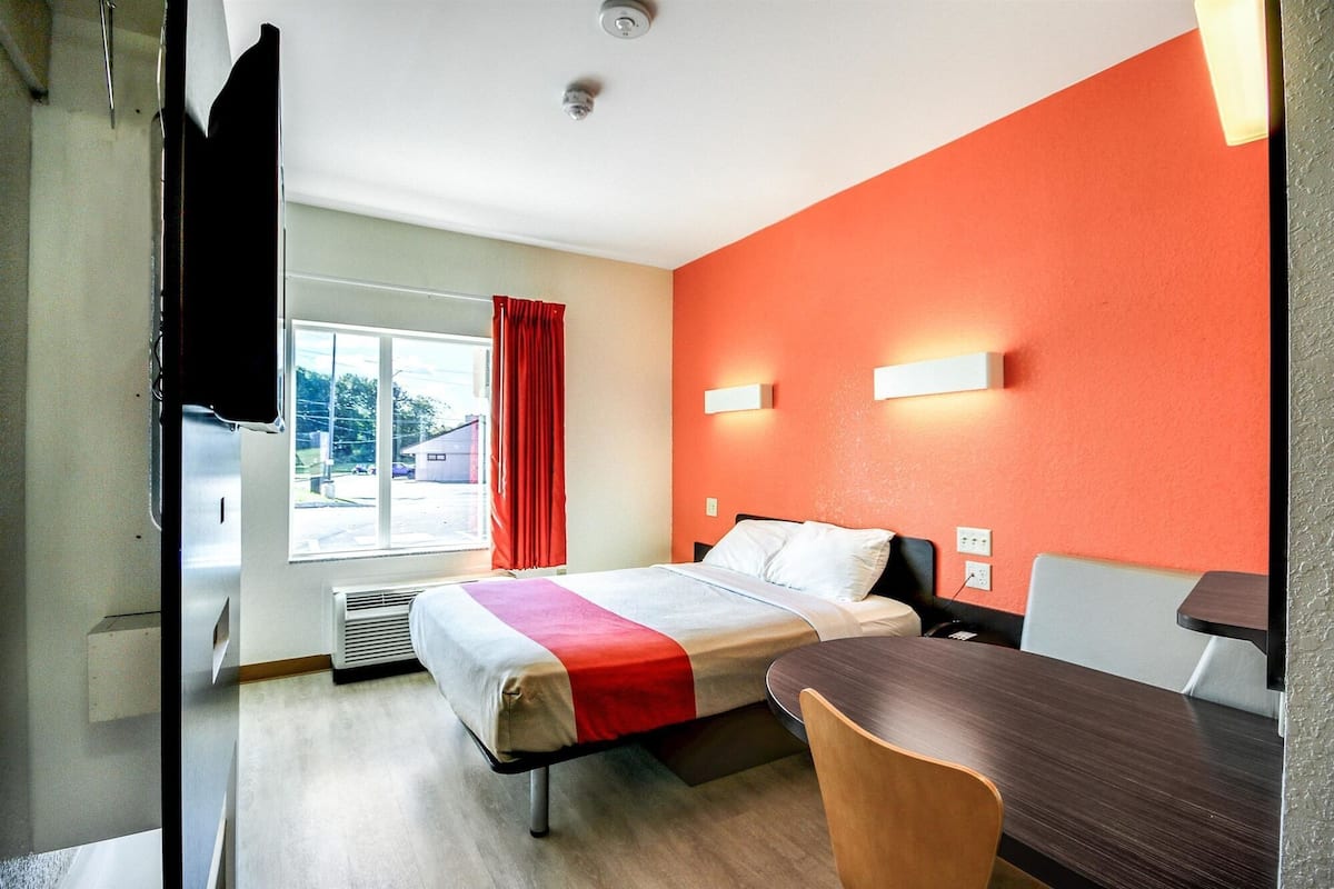 A hotel room with a bed, a desk, a chair, a window with a view of the outside, and a red wall.