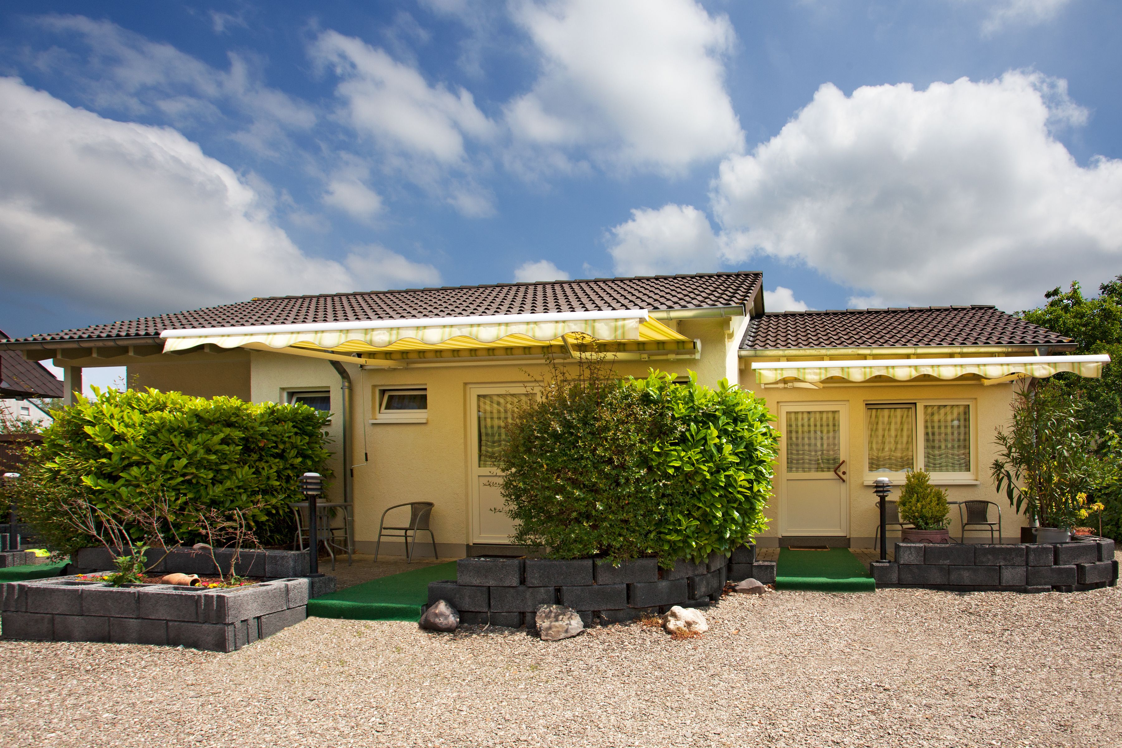 Une maison à deux étages, avec un toit recouvert de tuiles, un porche à l’avant surmonté d’un auvent jaune et un tapis vert menant à la porte. On y trouve des plantes en pot et un petit jardin agrémenté de gravier.