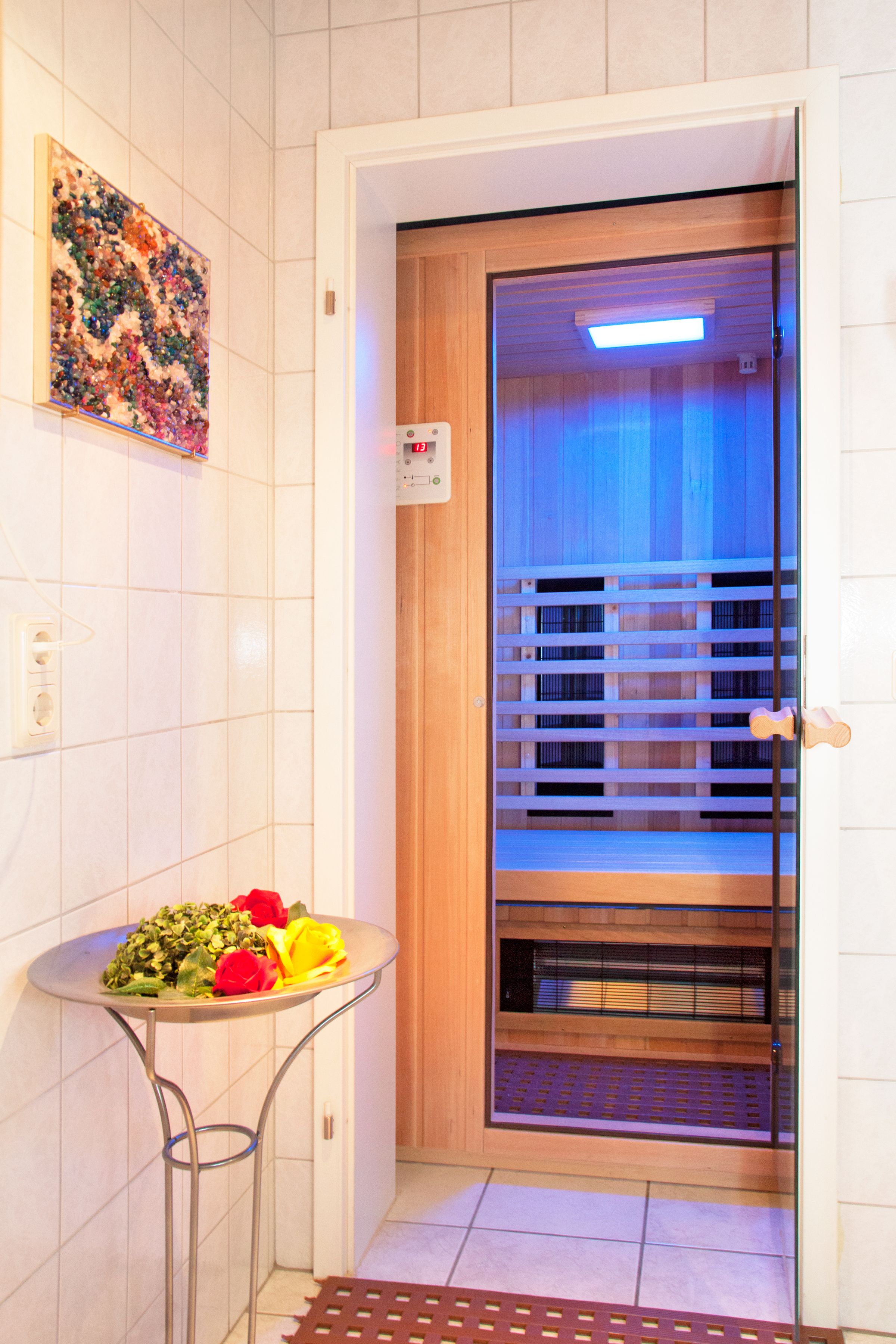 A bathroom with a tiled wall, a wooden door, a small table with a floral arrangement, and a shower area with a glass door.