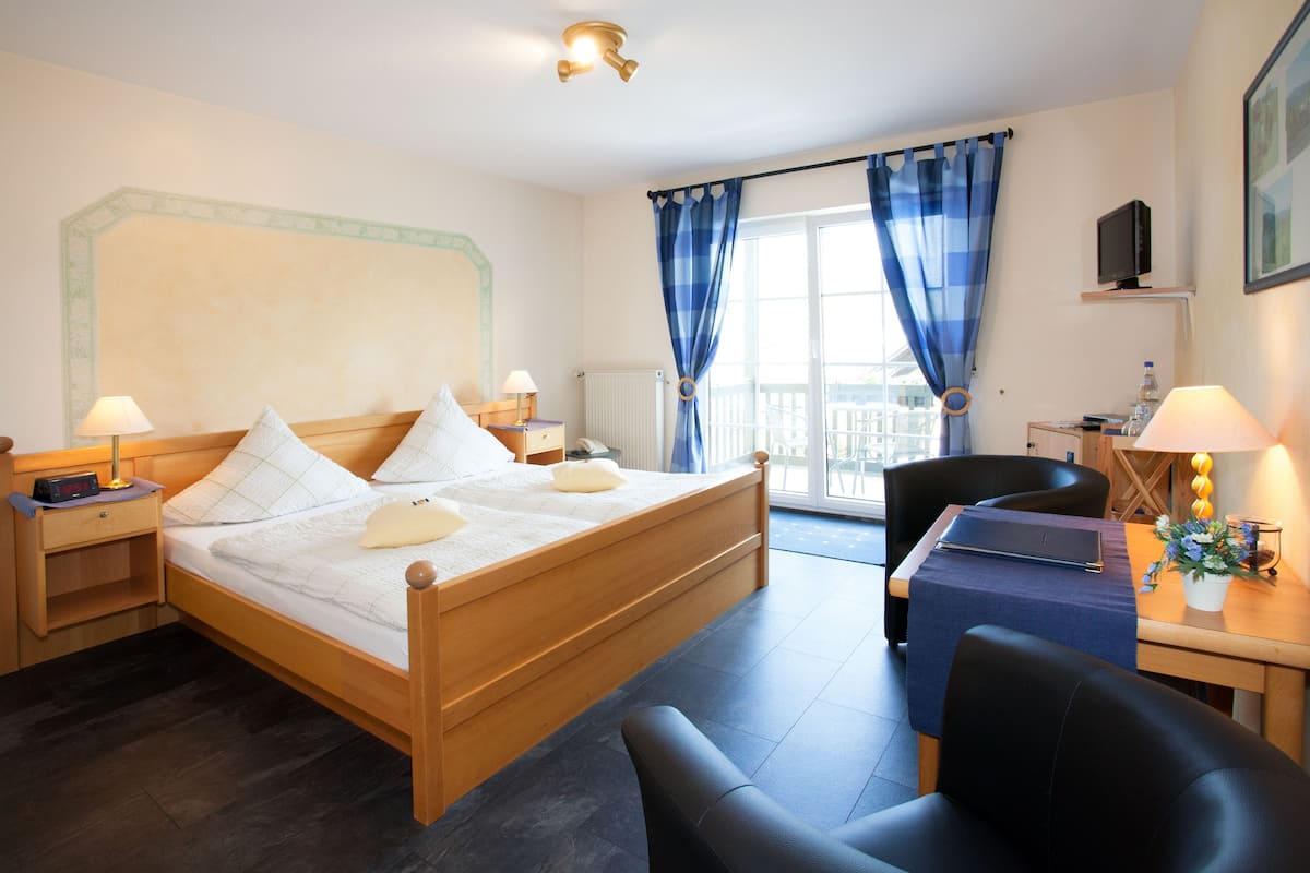 Une chambre d’hôtel avec un lit en bois, un bureau avec une chaise, une petite table avec une lampe, et une vue sur l’extérieur par une fenêtre ornée de rideaux bleus.