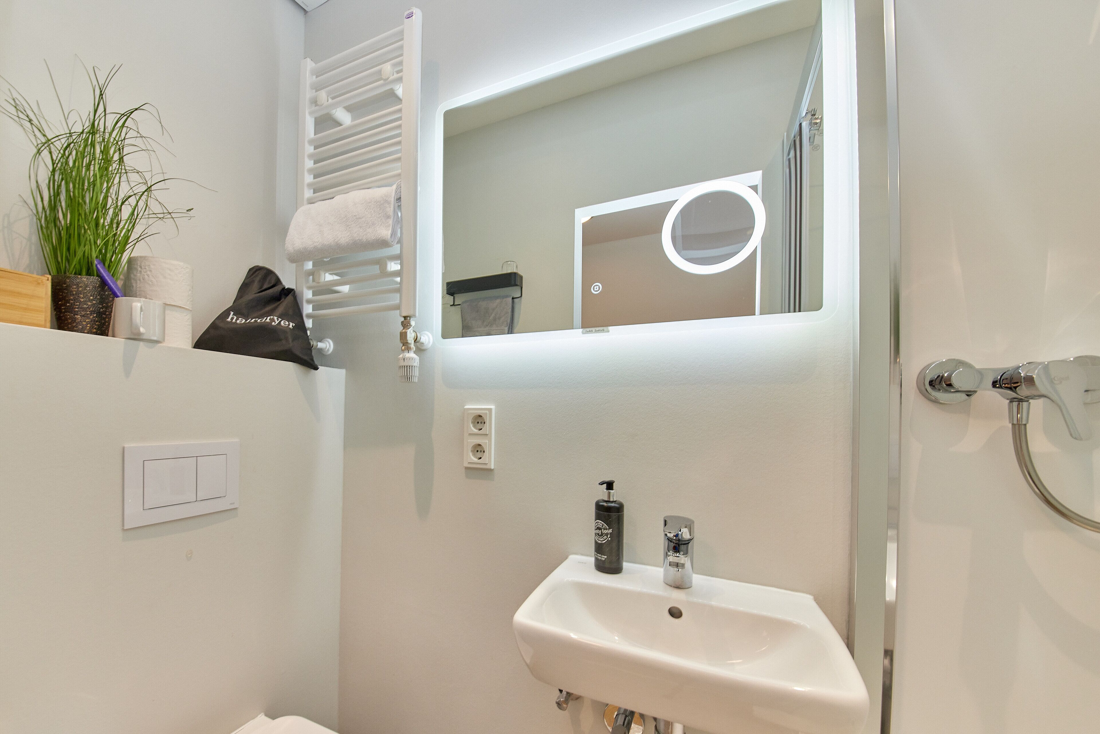 Un baño con lavamanos blanco, un espejo con luz, un perchero y una planta en maceta.
