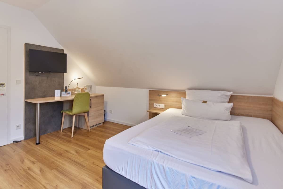 Une chambre d’hôtel moderne avec un plancher en bois, un bureau avec une chaise, un téléviseur à écran plat fixé au mur et un lit avec du linge de lit blanc.