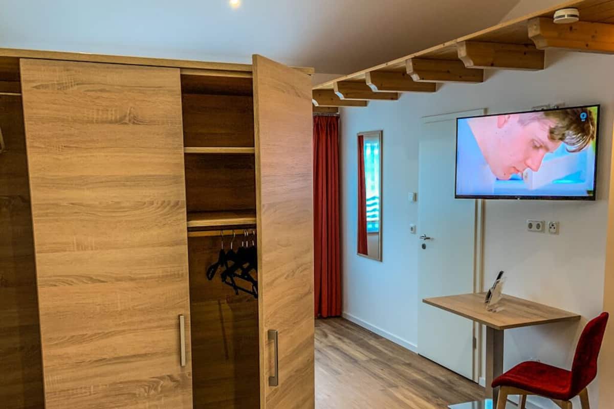 Une pièce comprenant une armoire en bois, une petite table avec une chaise, un téléviseur fixé au mur et un lit avec du linge de lit blanc.