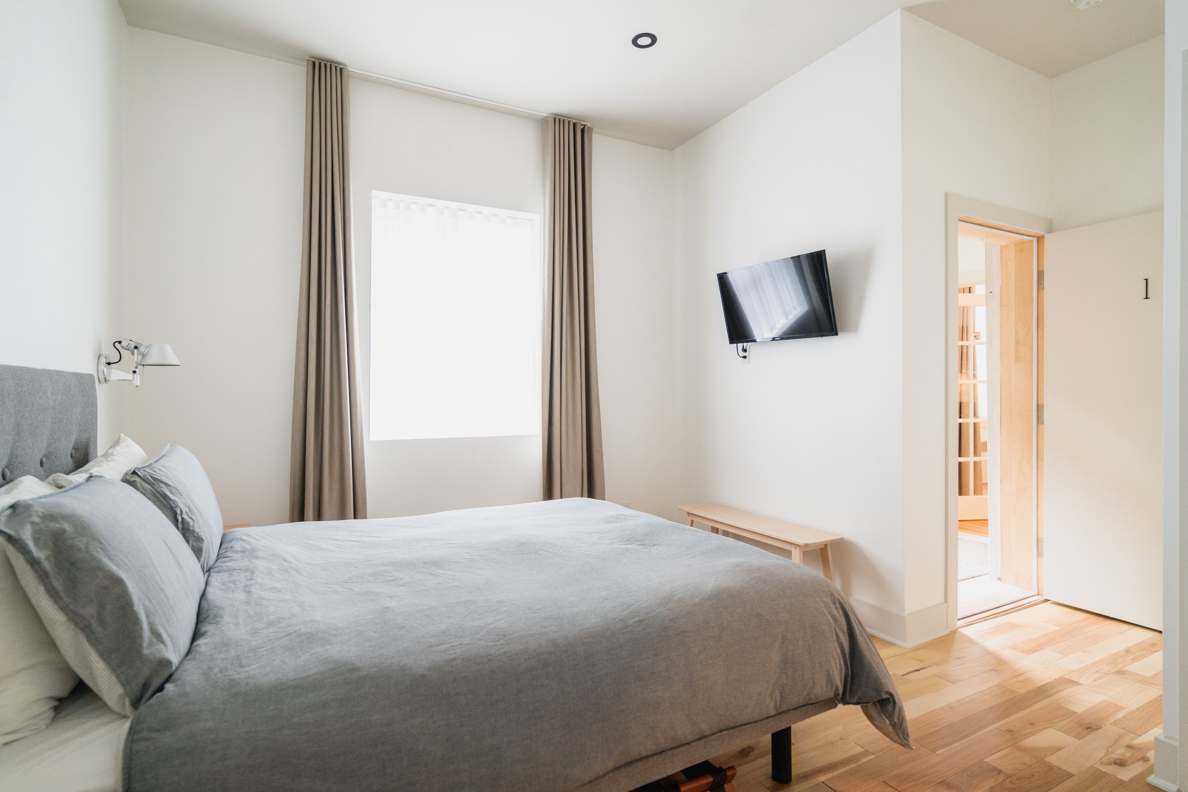 Ein Schlafzimmer mit Bett, Wandfernseher, Nachttisch und einem Fenster mit Vorhängen.