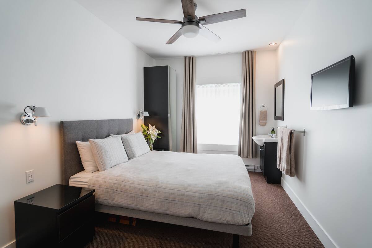 Habitación de hotel moderna con cama, mesita de noche, ventilador de techo, ventana con cortinas y televisor montado en la pared.