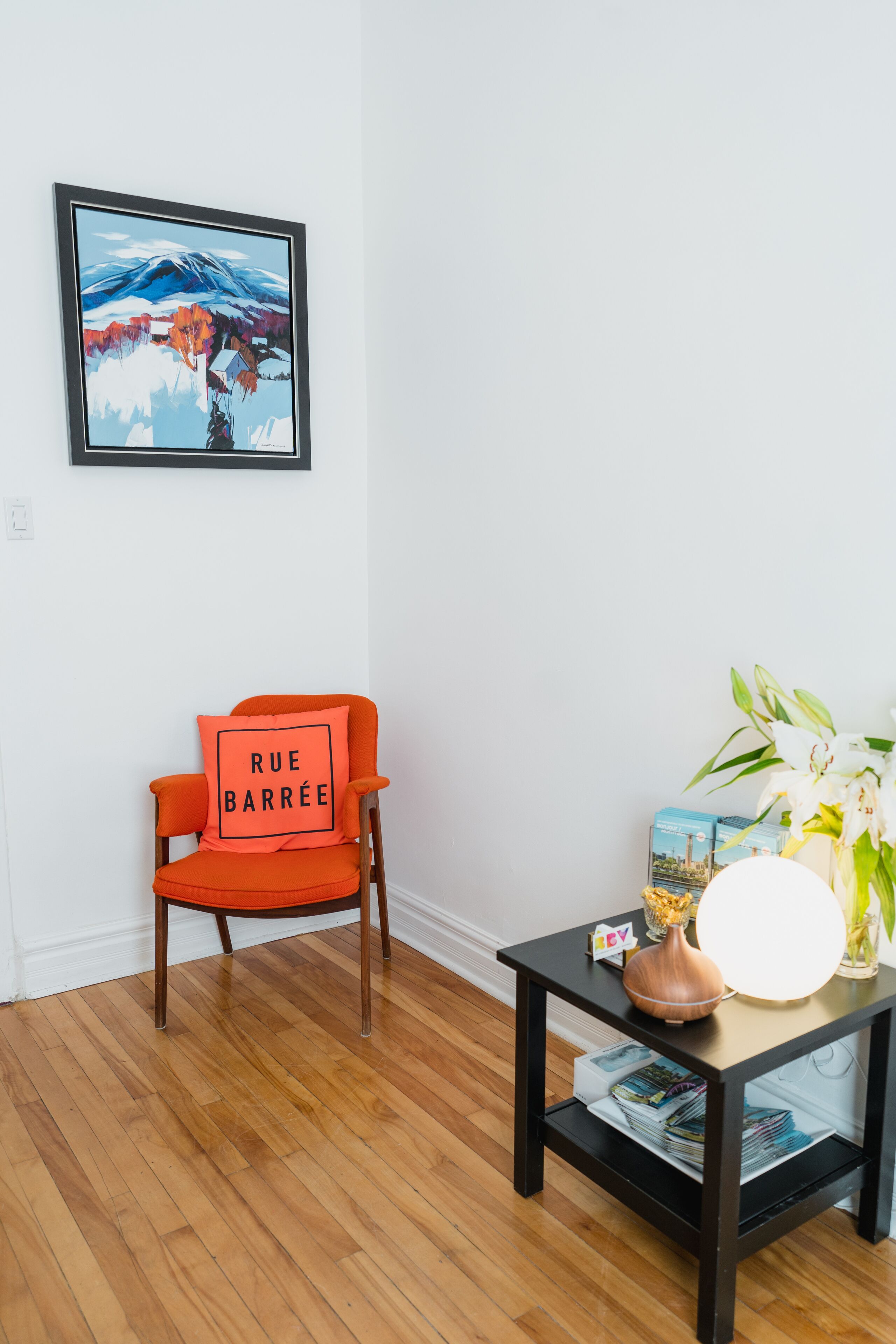 A room with a wooden floor, a red chair with a cushion labeled "RUE BARRÉE," a black table with a vase of flowers, a framed picture on the wall, and a stack of magazines on the table.