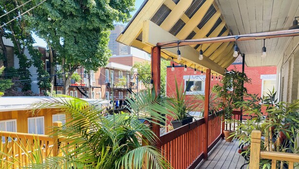 Una terraza de madera con plantas en macetas, barandilla de madera y vista de edificios al fondo.