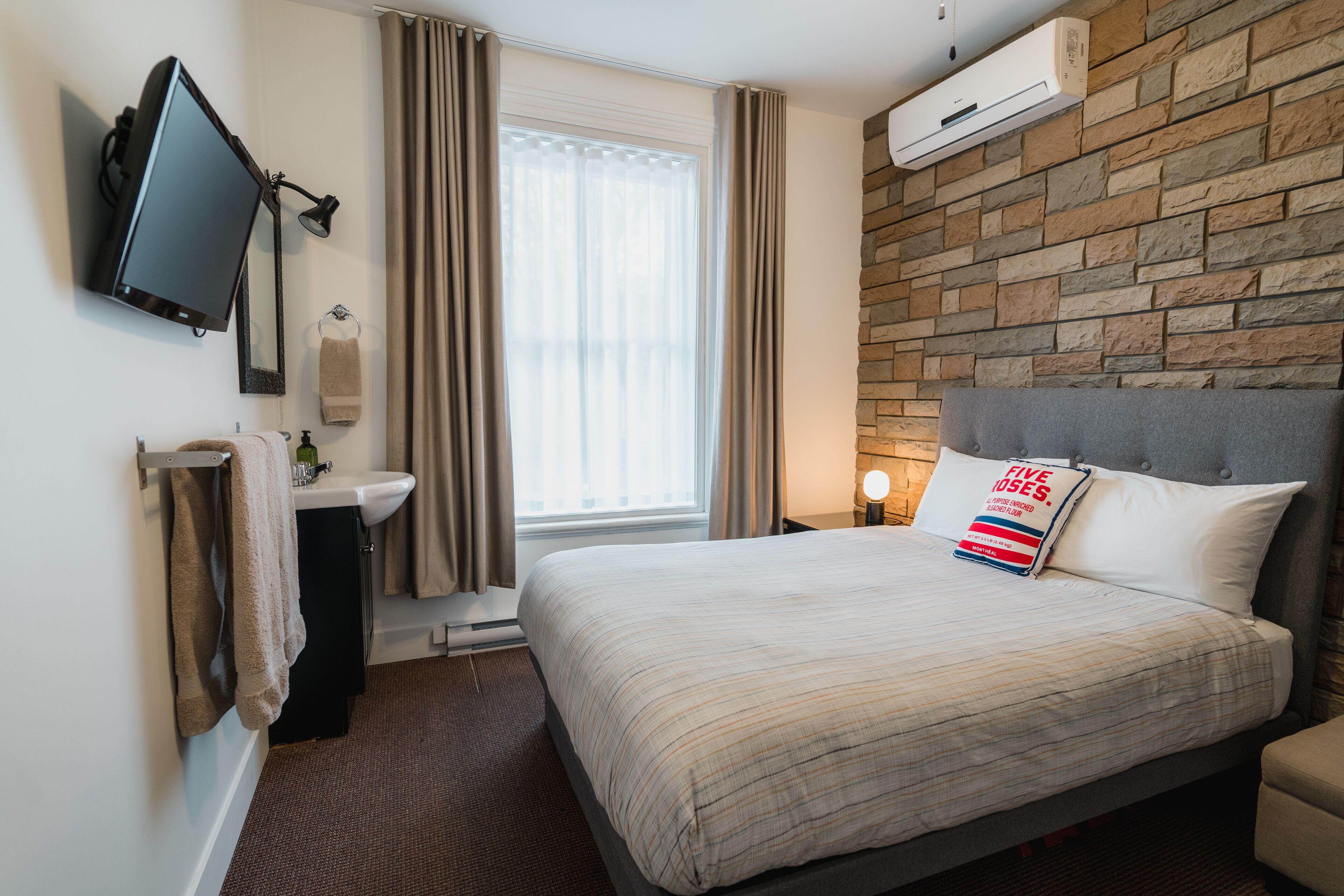 Une chambre d’hôtel avec un lit, un téléviseur fixé au mur, une salle de bain avec un lavabo et un miroir, et un mur en pierre.