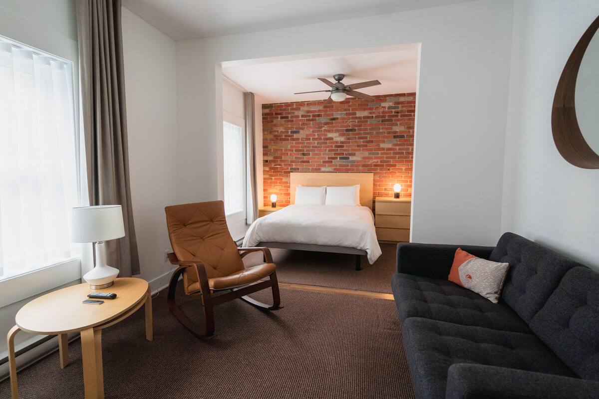 Habitación de hotel con pared decorada en ladrillo, cama con ropa blanca, sofá gris oscuro, silla de madera, mesita redonda con lámpara y ventilador de techo.