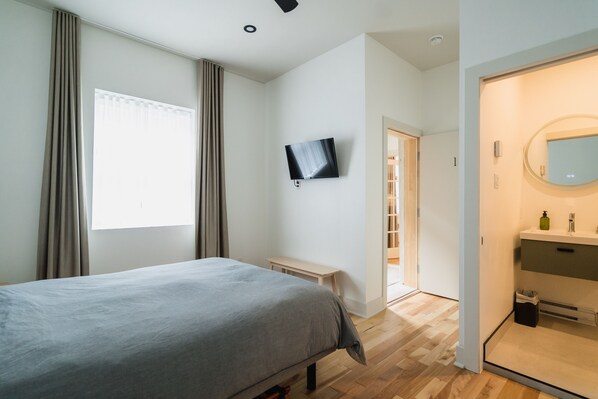 Un dormitorio con cama, televisión montada en la pared, una mesita de noche pequeña y un baño con espejo y lavabo.
