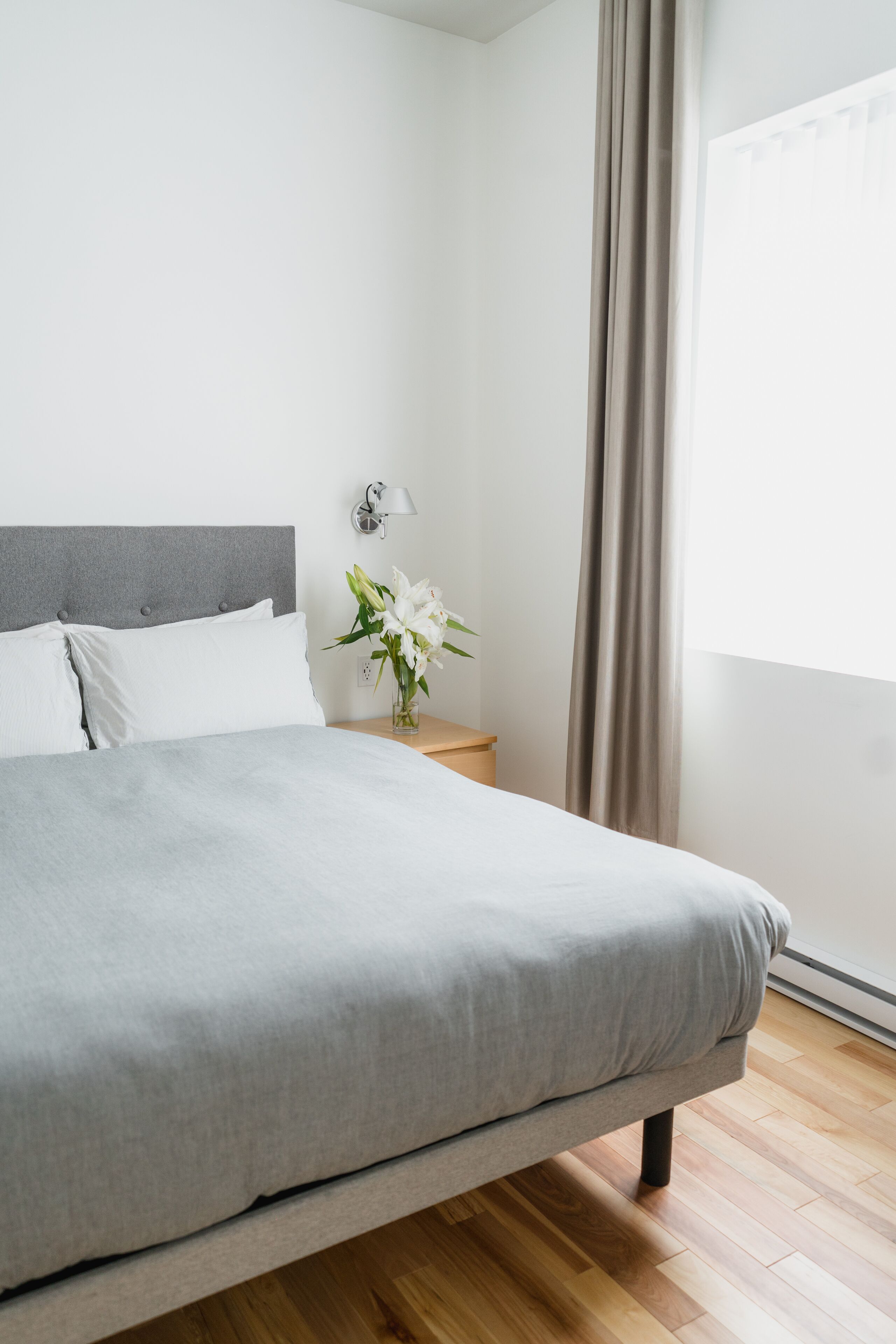Una cama bien tendida con cabecera gris y almohadas blancas, una mesita de noche con una lámpara y un florero, y una ventana con cortinas.