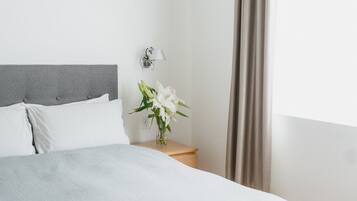 Una cama bien tendida con cabecera gris y almohadas blancas, una mesita de noche con una lámpara y un florero, y una ventana con cortinas.