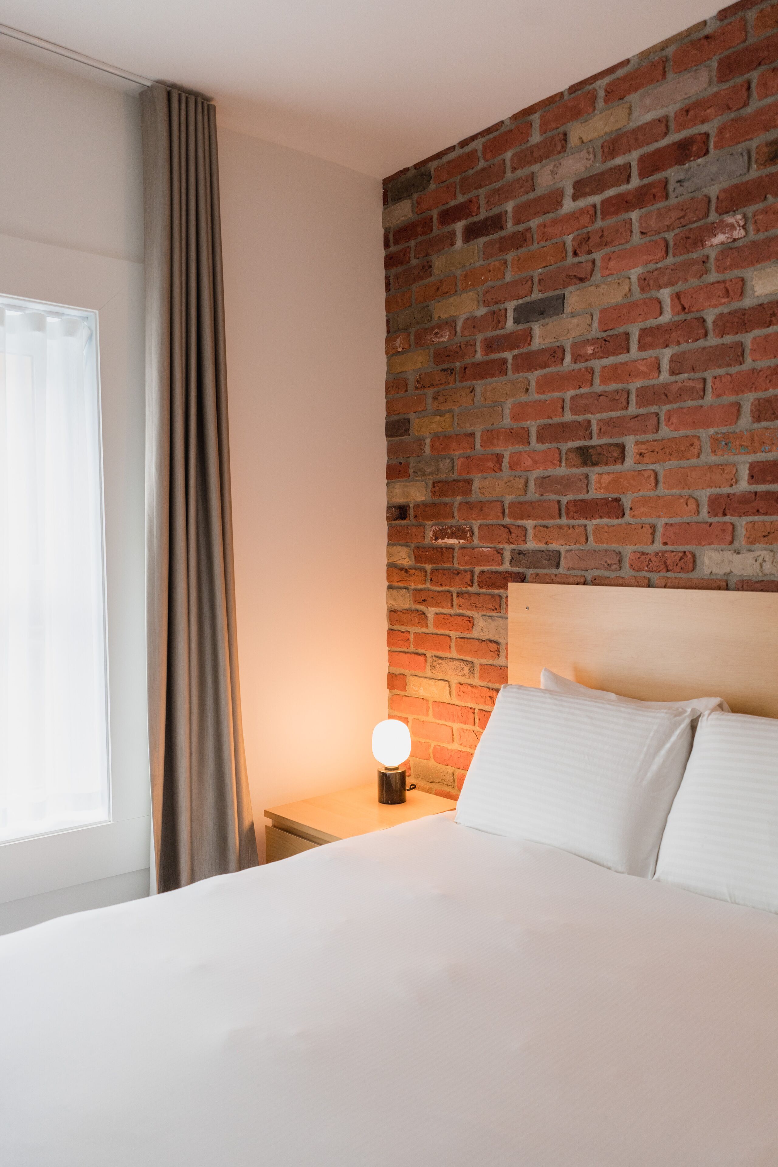 Una camera d'albergo con una parete in mattoni, un letto con lenzuola bianche, un comodino con una lampada e una finestra con tende.