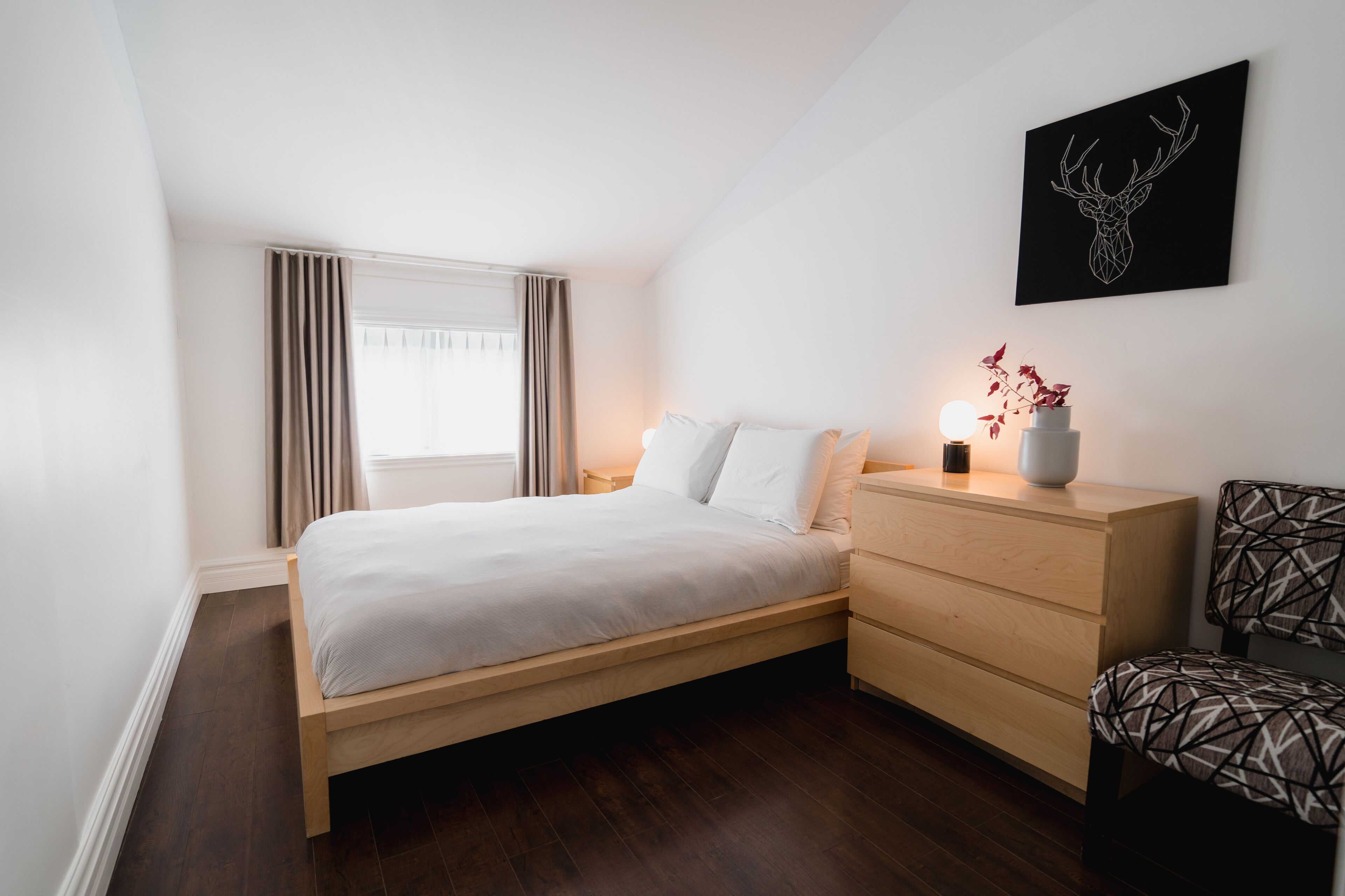A modern bedroom with a wooden bed, a nightstand, a dresser, and a wall-mounted deer head artwork.