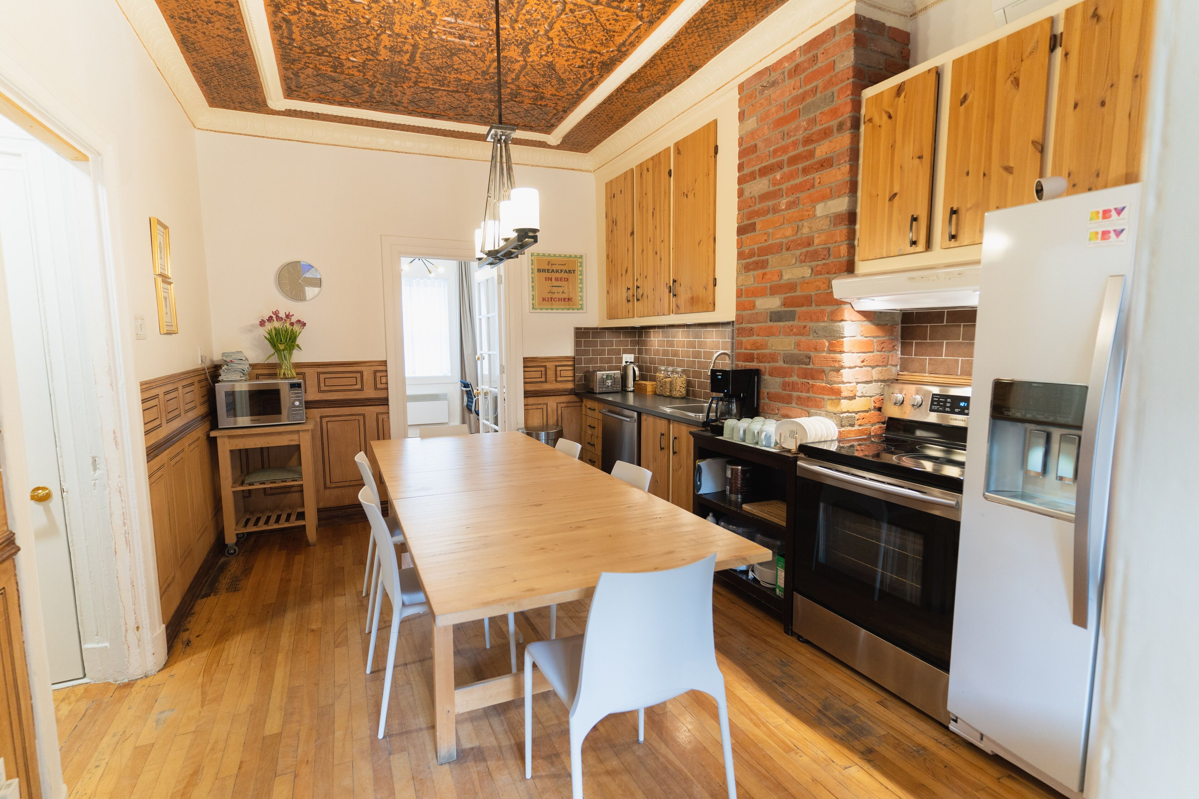 A kitchen with wooden cabinets, a large wooden table, white chairs, a refrigerator, and a brick wall.