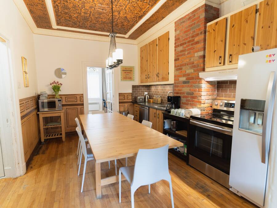 A kitchen with wooden cabinets, a large wooden table, white chairs, a refrigerator, and a brick wall.