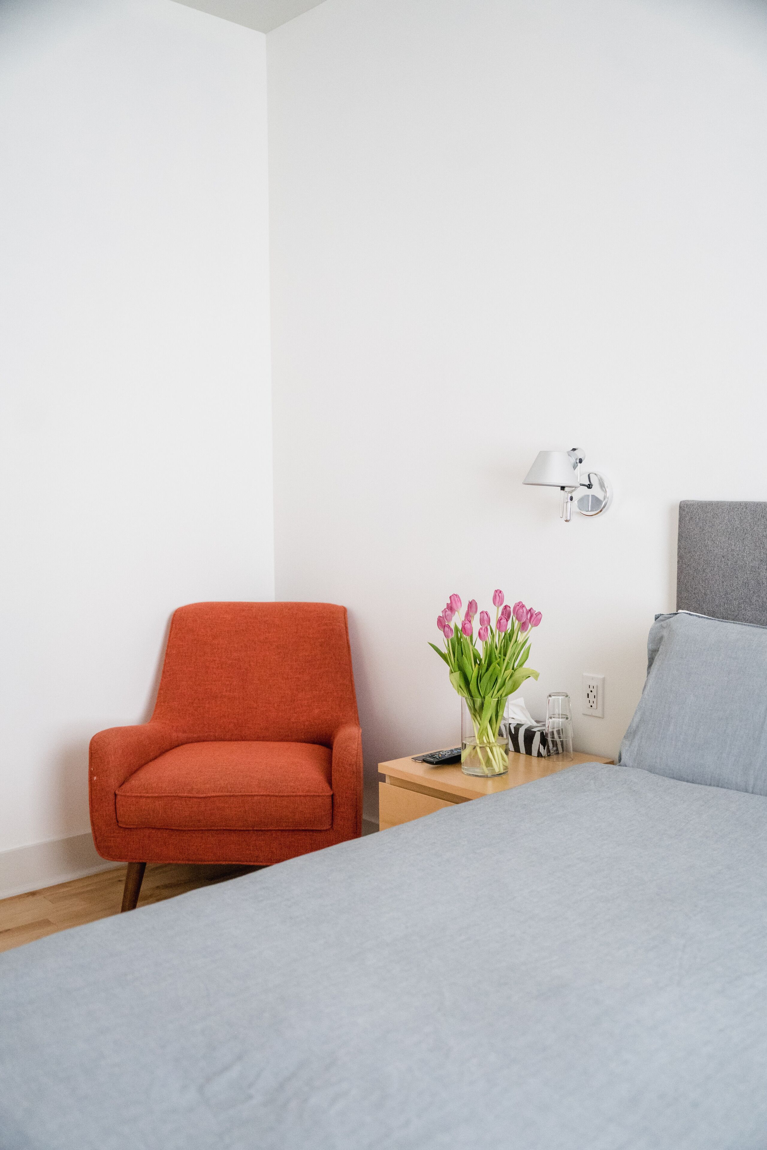 A bedroom with a bed, a chair, a nightstand with a lamp, and a vase with flowers.