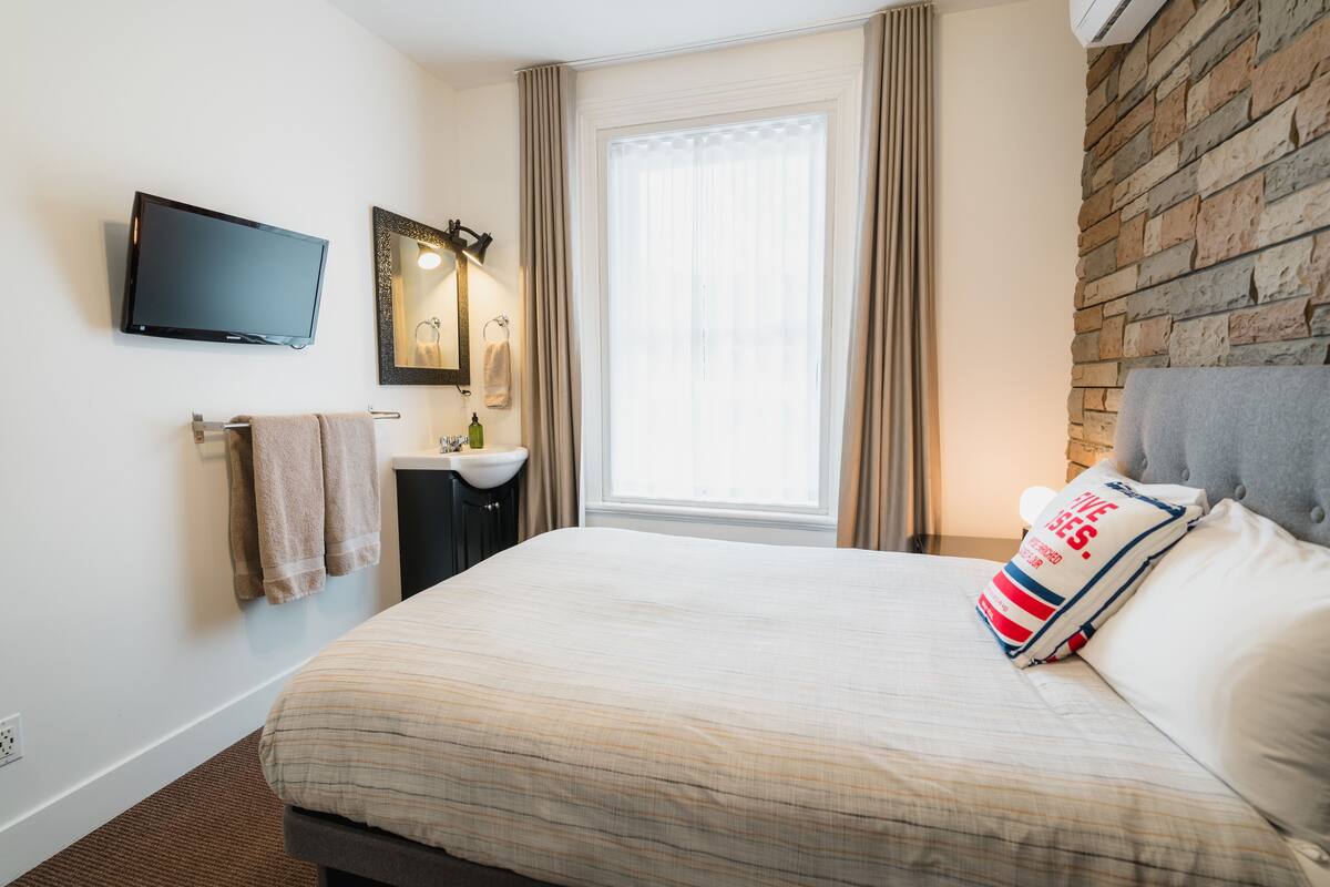 Habitación de hotel con cama, televisor de pantalla plana, espejo, ventana con cortinas, lavabo y pared de piedra.