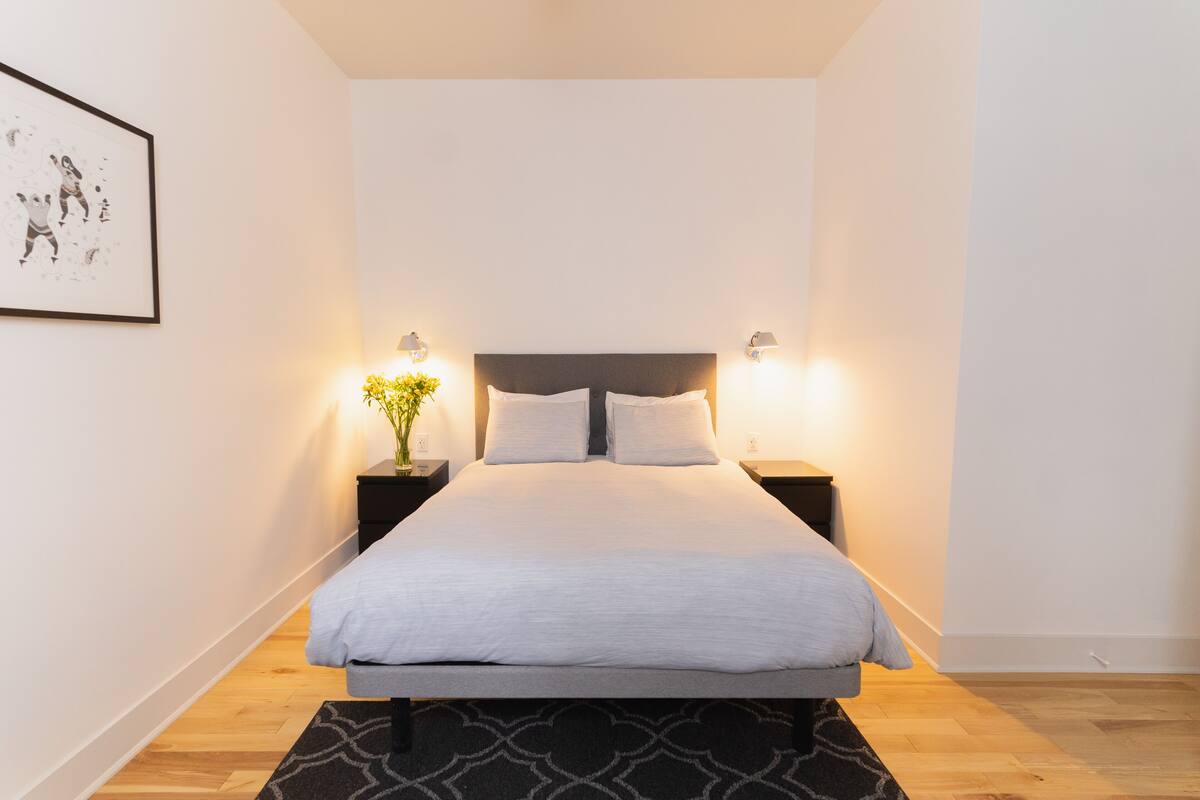 Una cama bien hecha con edredón gris, dos mesitas de noche con lámparas y un cuadro en la pared.