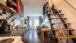 Una cocina compacta con estufa, cafetera y armarios de madera. Un área de comedor con mesa y sillas está al lado de una escalera.
