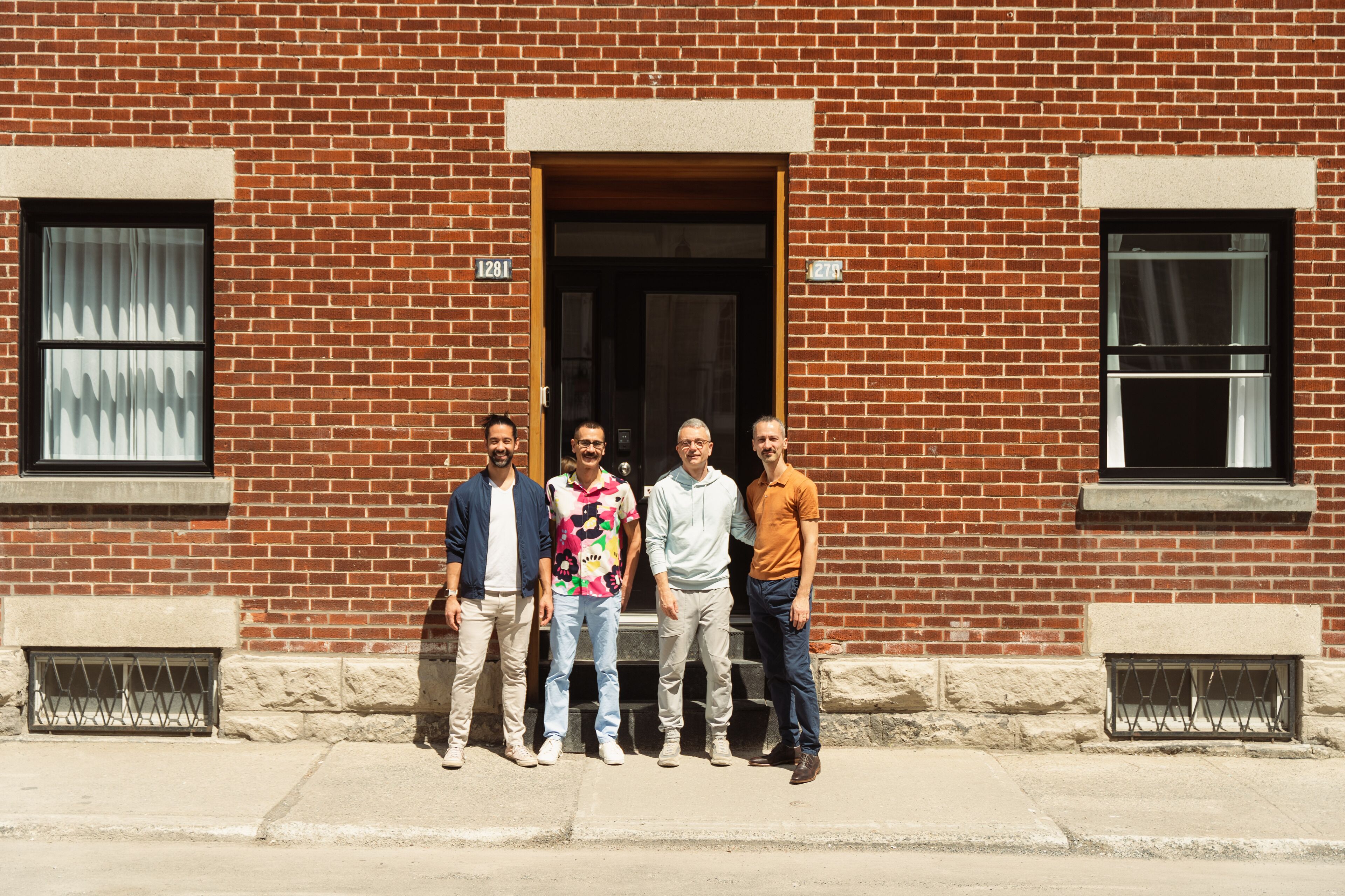 A group of five men standing in front of a brick building with numbered windows and a door.