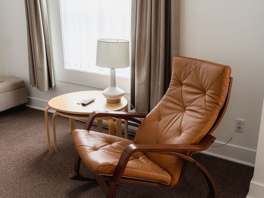 Ein Zimmer mit braunem Ledersessel, einem kleinen runden Tisch mit Lampe und einem Fenster mit Vorhängen.