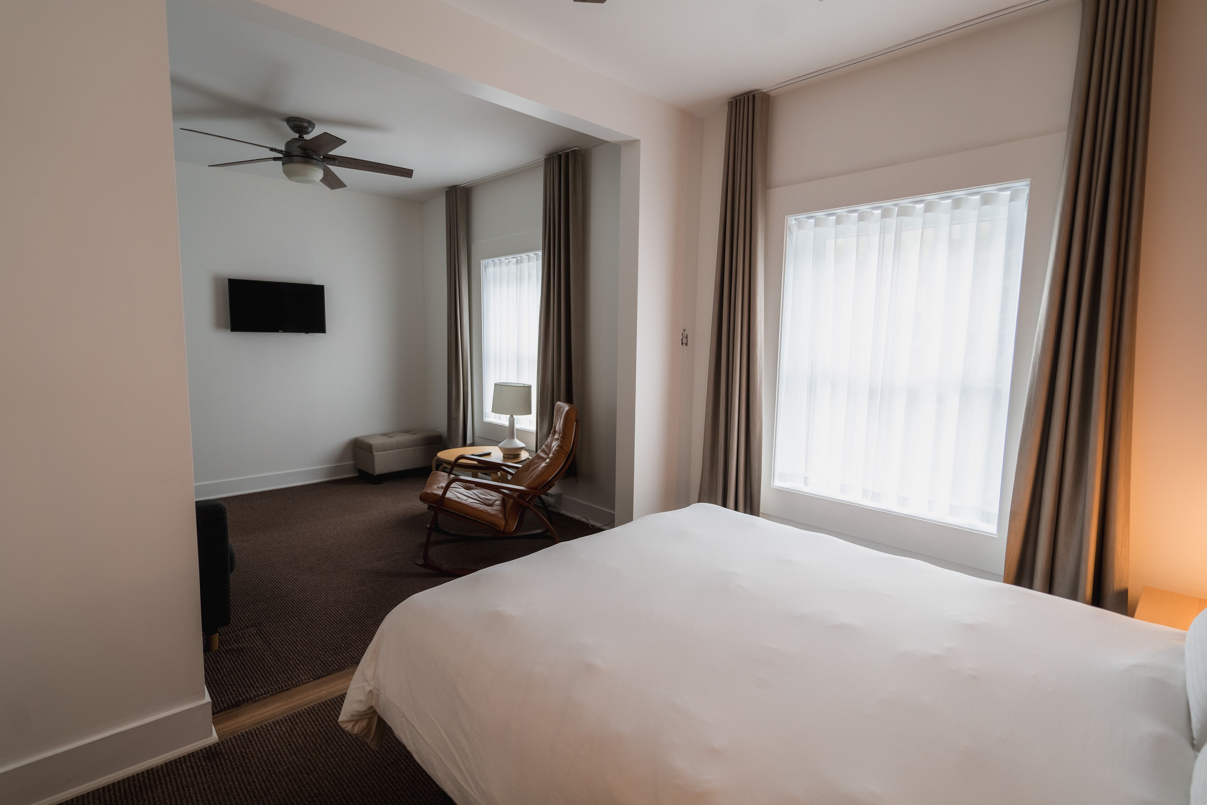A hotel room with a large bed, a chair, a desk, a ceiling fan, and a window with curtains.
