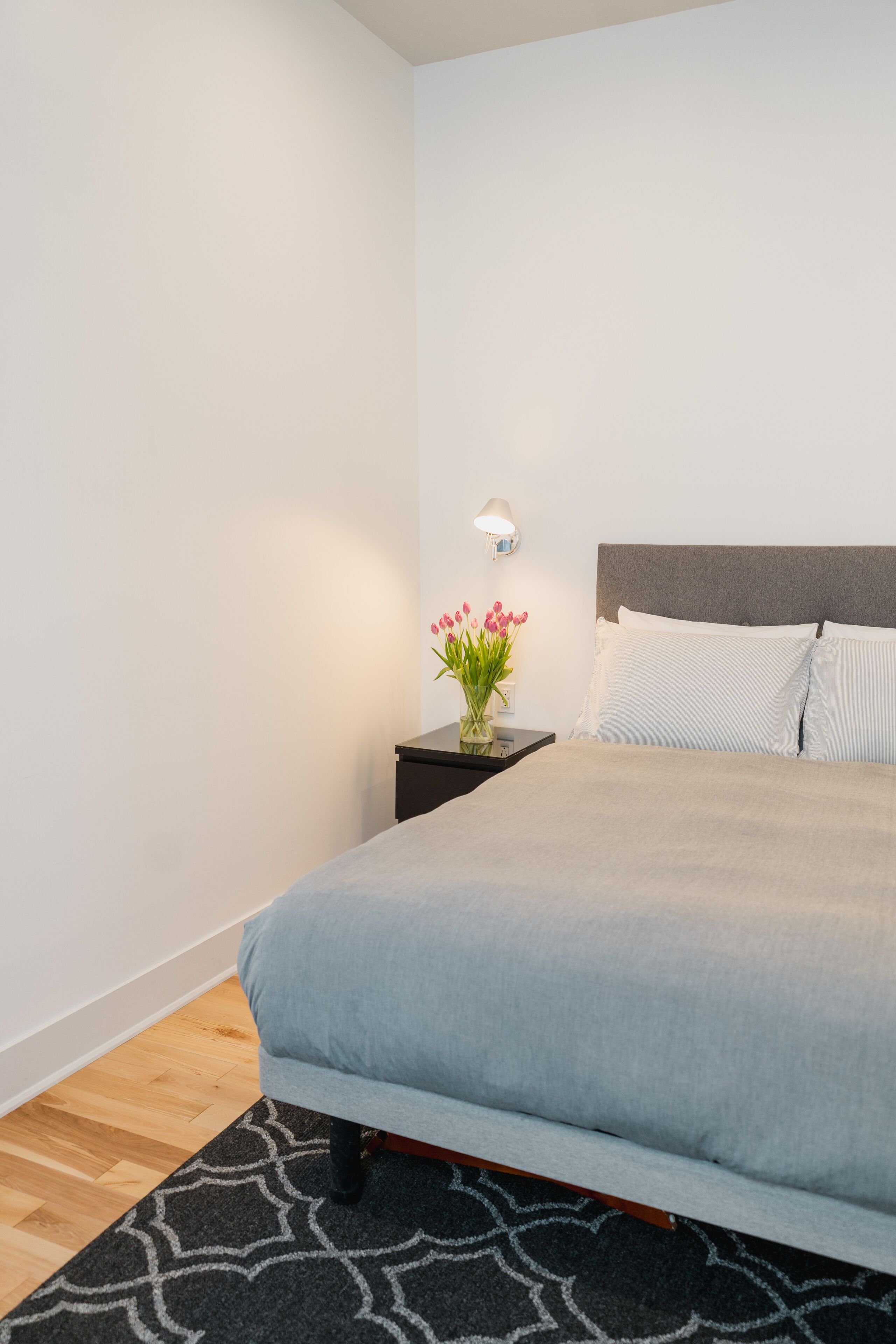 Una cama bien tendida con un cubrecolcha gris, una mesita de noche con una lámpara y un florero con flores.