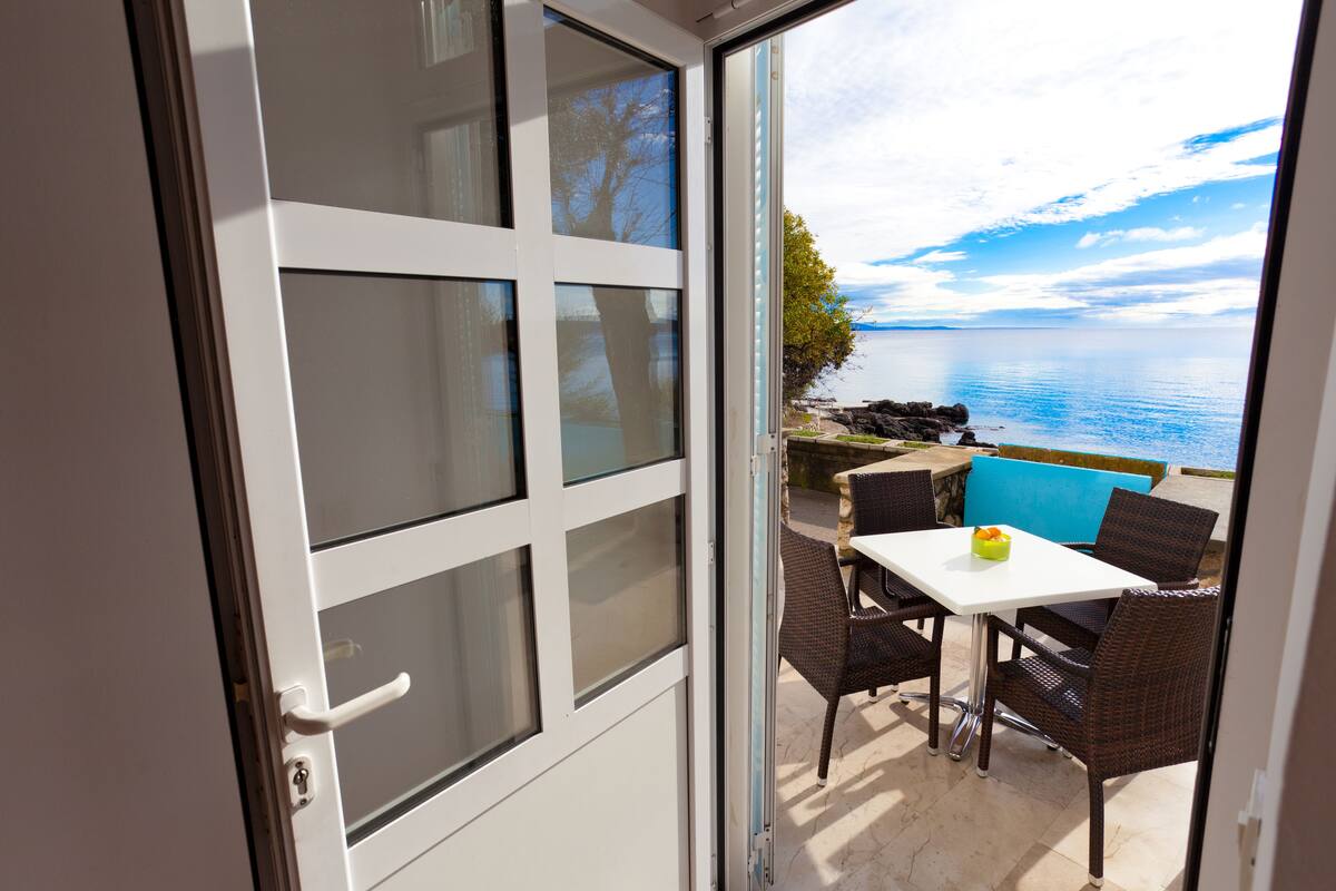 A room with a sliding glass door leading to an outdoor seating area with a table and chairs, overlooking a body of water.