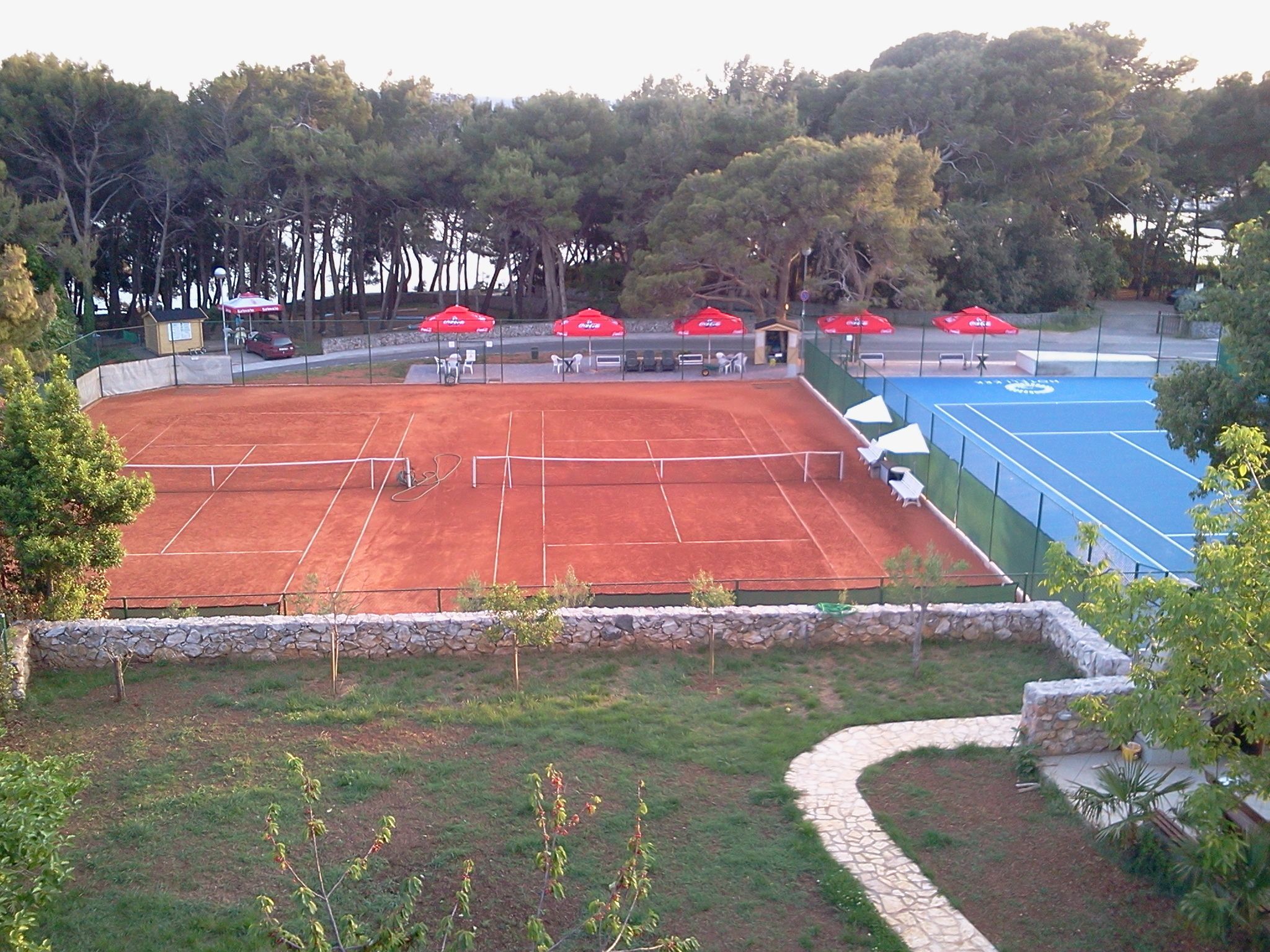 A tennis court with a stone wall and a blue area, surrounded by trees and outdoor seating.