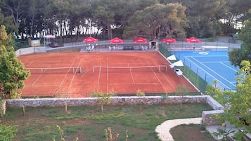 A tennis court with a stone wall and a blue area, surrounded by trees and outdoor seating.