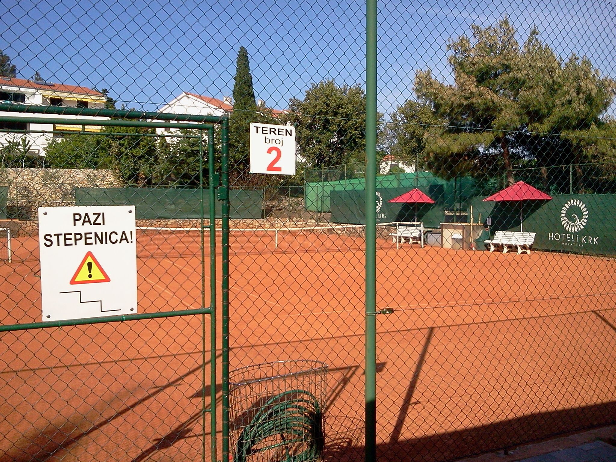 A tennis court with a warning sign, a bench, and a building in the background.