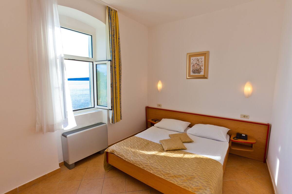 A hotel room with a bed, a window with curtains, a bedside table, a wall-mounted light, and a framed picture.