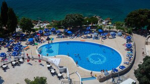 A large, circular swimming pool surrounded by lounge chairs and umbrellas, with a clear view of the sea in the background.