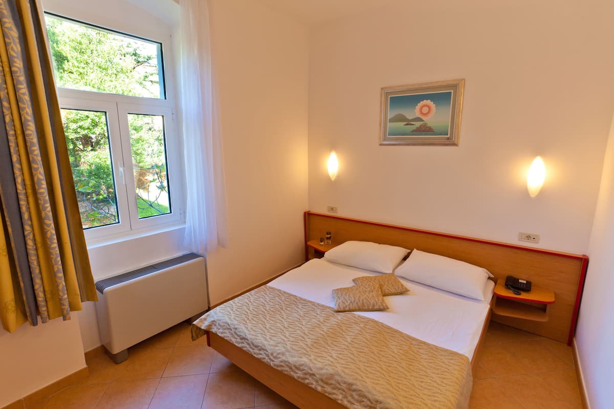 A bedroom with a bed, bedside tables, a nightstand, a window with curtains, a radiator, and a painting on the wall.