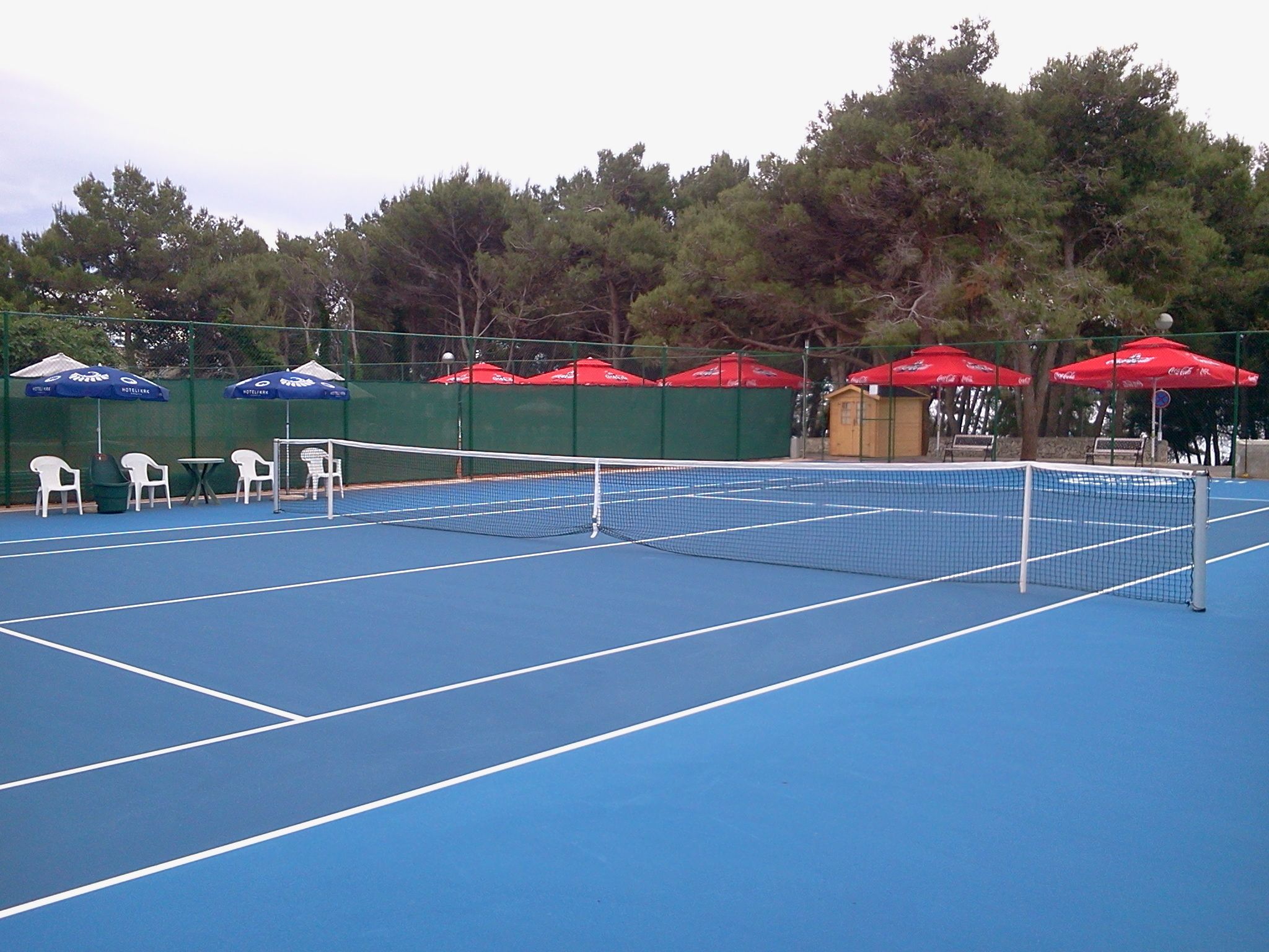 A tennis court with a net, surrounded by green fencing and red umbrellas.