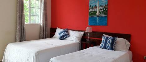 A room with two beds, a window with curtains, a lamp, and a painting on the wall.