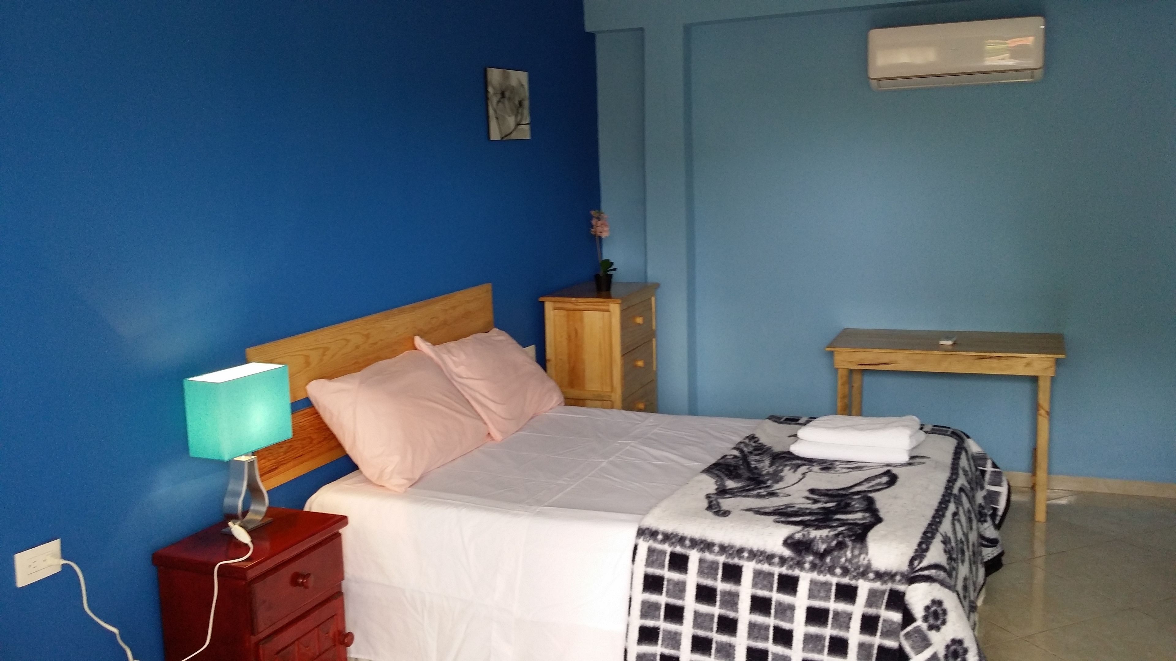 A bedroom with a bed, a nightstand, a dresser, a small table, and an air conditioning unit.