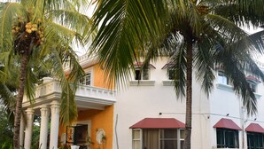 Un edificio de dos pisos con un balcón, un toldo rojo y una palmera en primer plano.