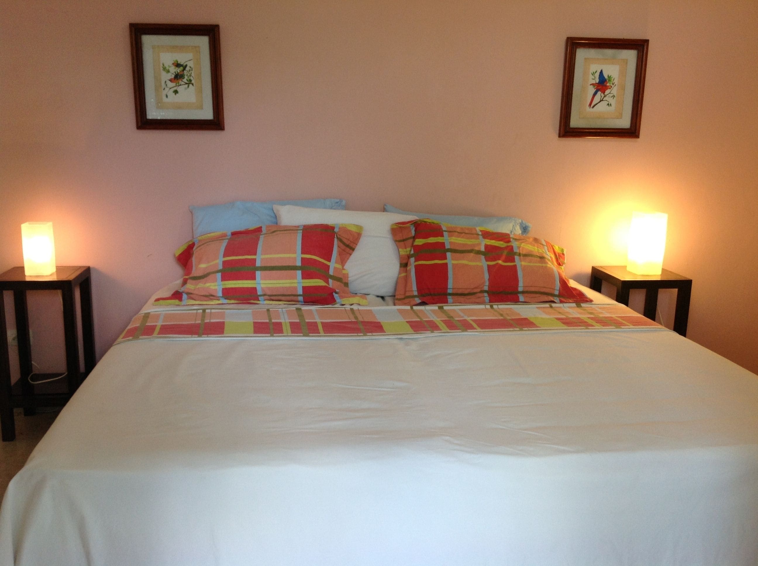 Una cama bien tendida con una colcha blanca y almohadas a rayas coloridas, flanqueada por dos mesitas de noche con lámparas, en una habitación con paredes rosa claro y un cuadro con motivos florales sobre la cama.