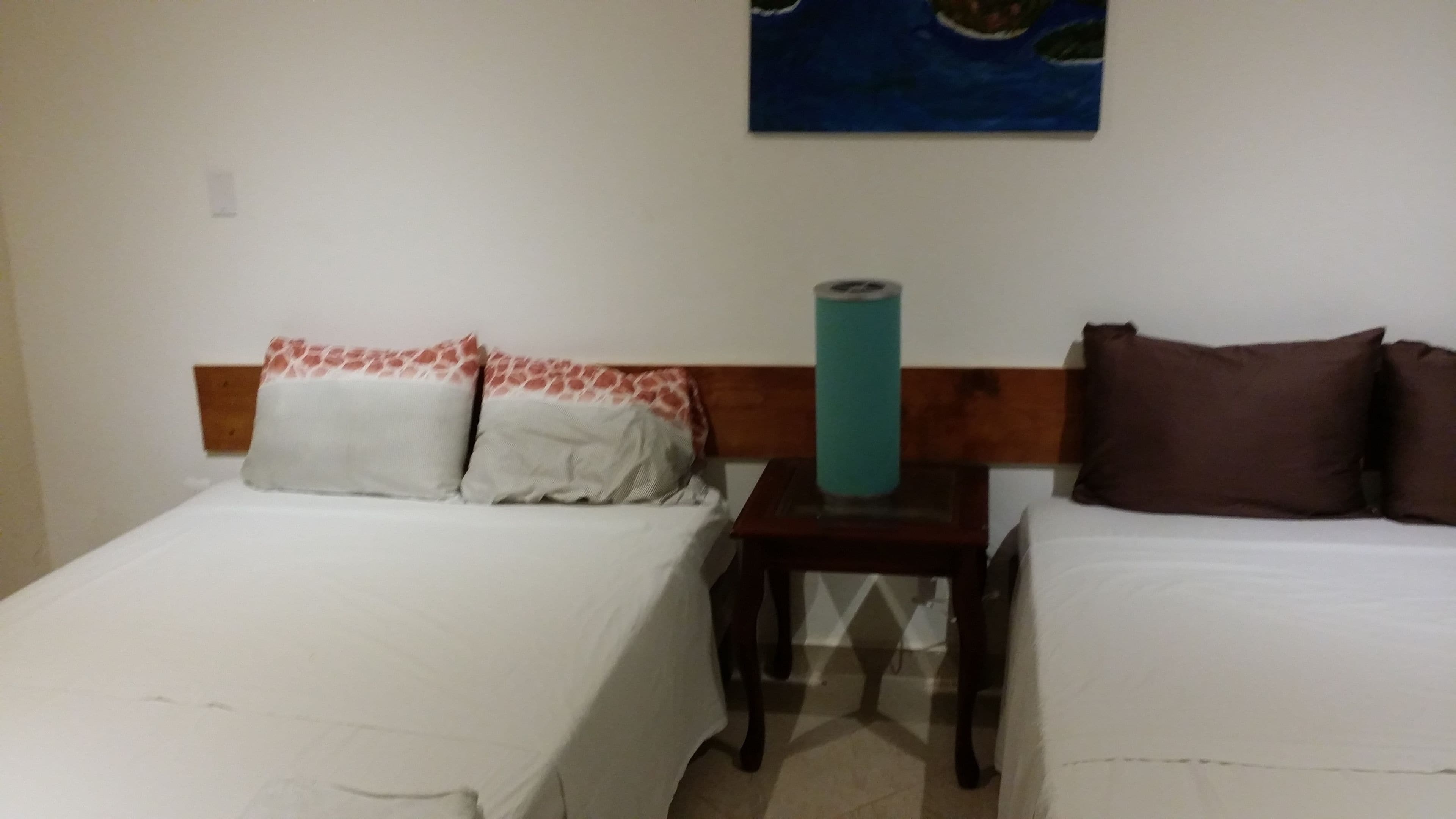 A hotel room with two single beds, a wooden headboard, a small table with a teal-colored object, and a painting on the wall.
