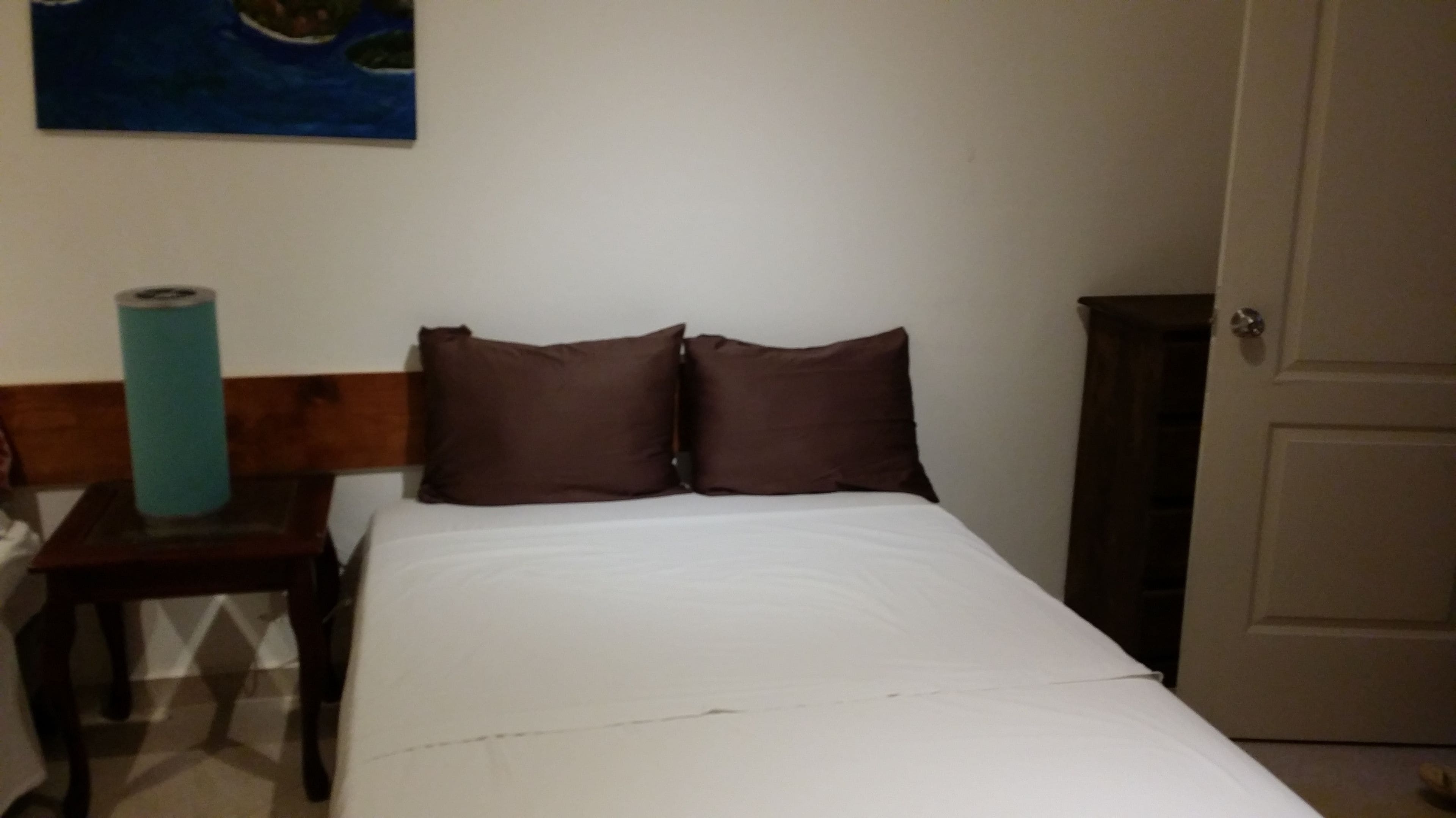 A bedroom with a bed, two pillows, a wooden headboard, a small table with a blue object, and a door.