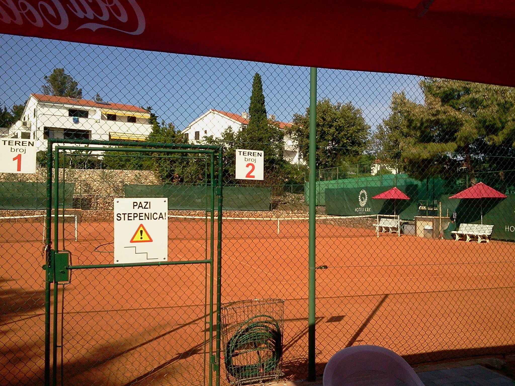 Un court de tennis avec un panneau d’avertissement, des courts numérotés et une banderole Coca-Cola.