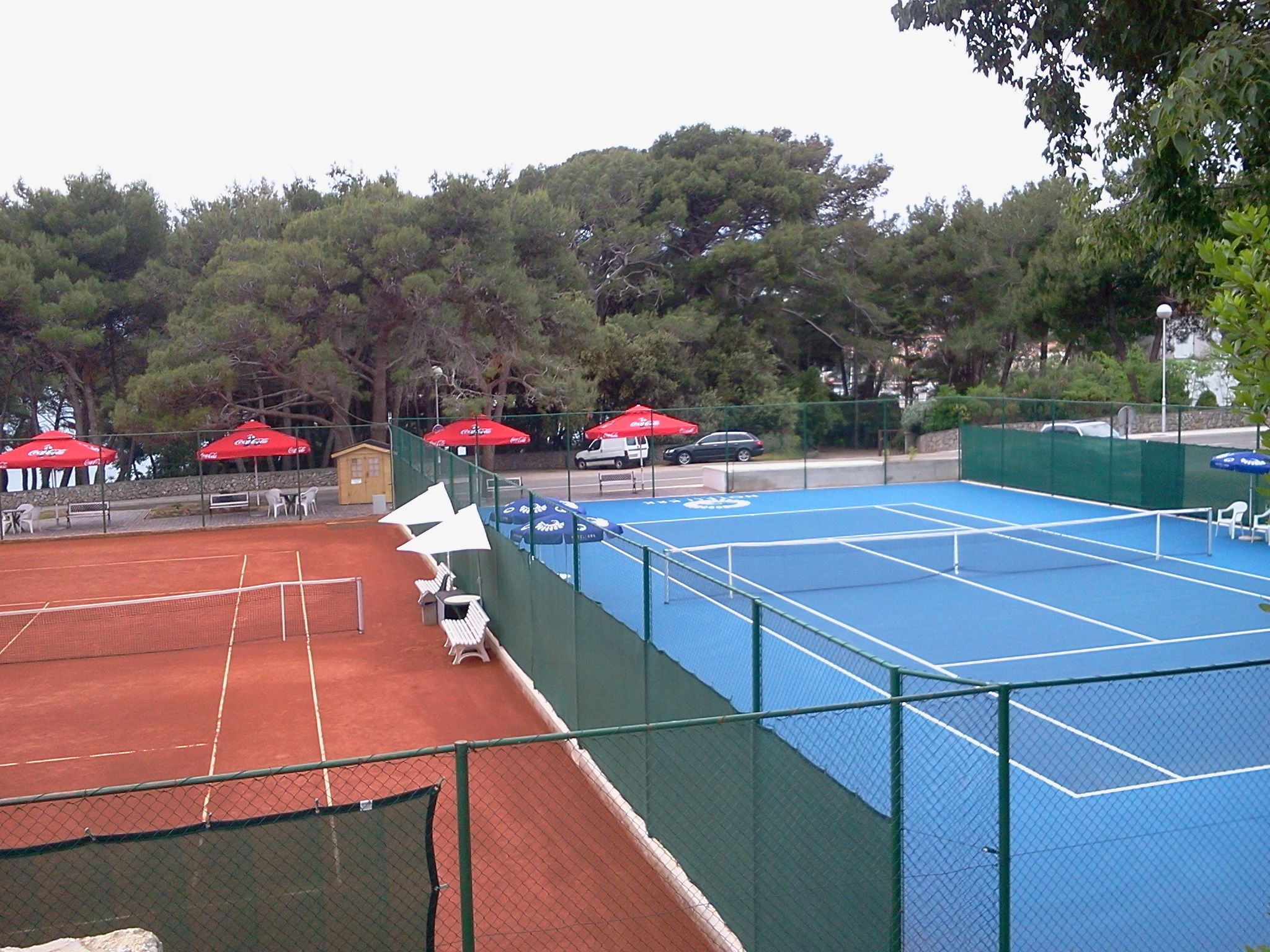 Ein Tennisplatz mit Netz, umgeben von grünem Zaun und roten Sonnenschirmen.