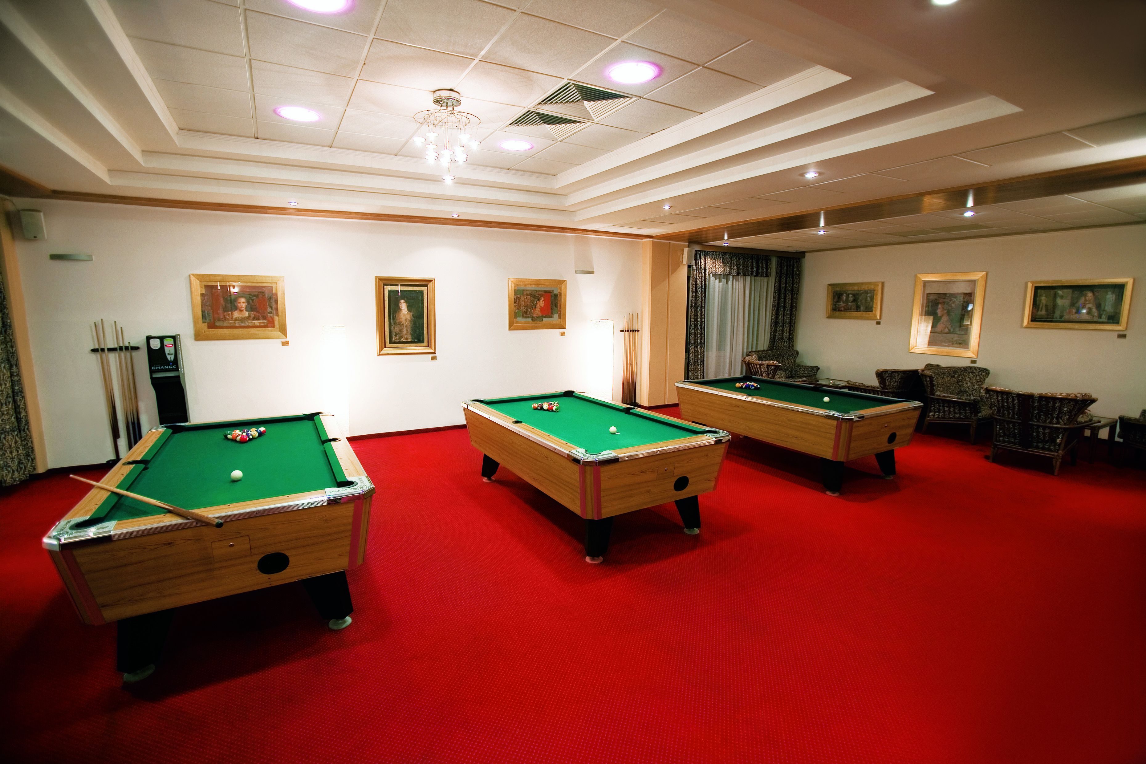 A pool room with three pool tables, red carpet flooring, and framed artwork on the walls.