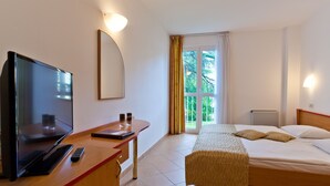 A hotel room with a bed, a desk with a TV, a remote, and a window with curtains. - Villa Lovorka - Hotel Resort Drazica (Krk)