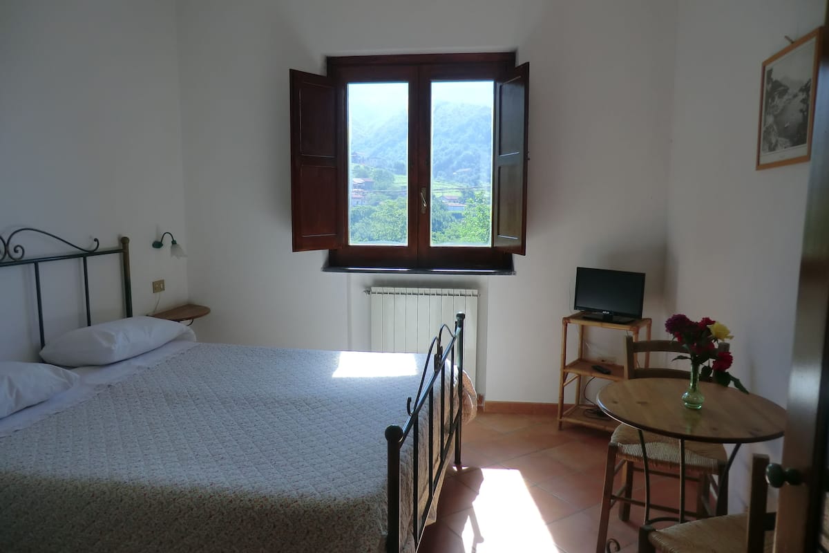 Een slaapkamer met een bed, een bureau met een computer, een stoel, een klein tafeltje met bloemen en een raam met open luiken.