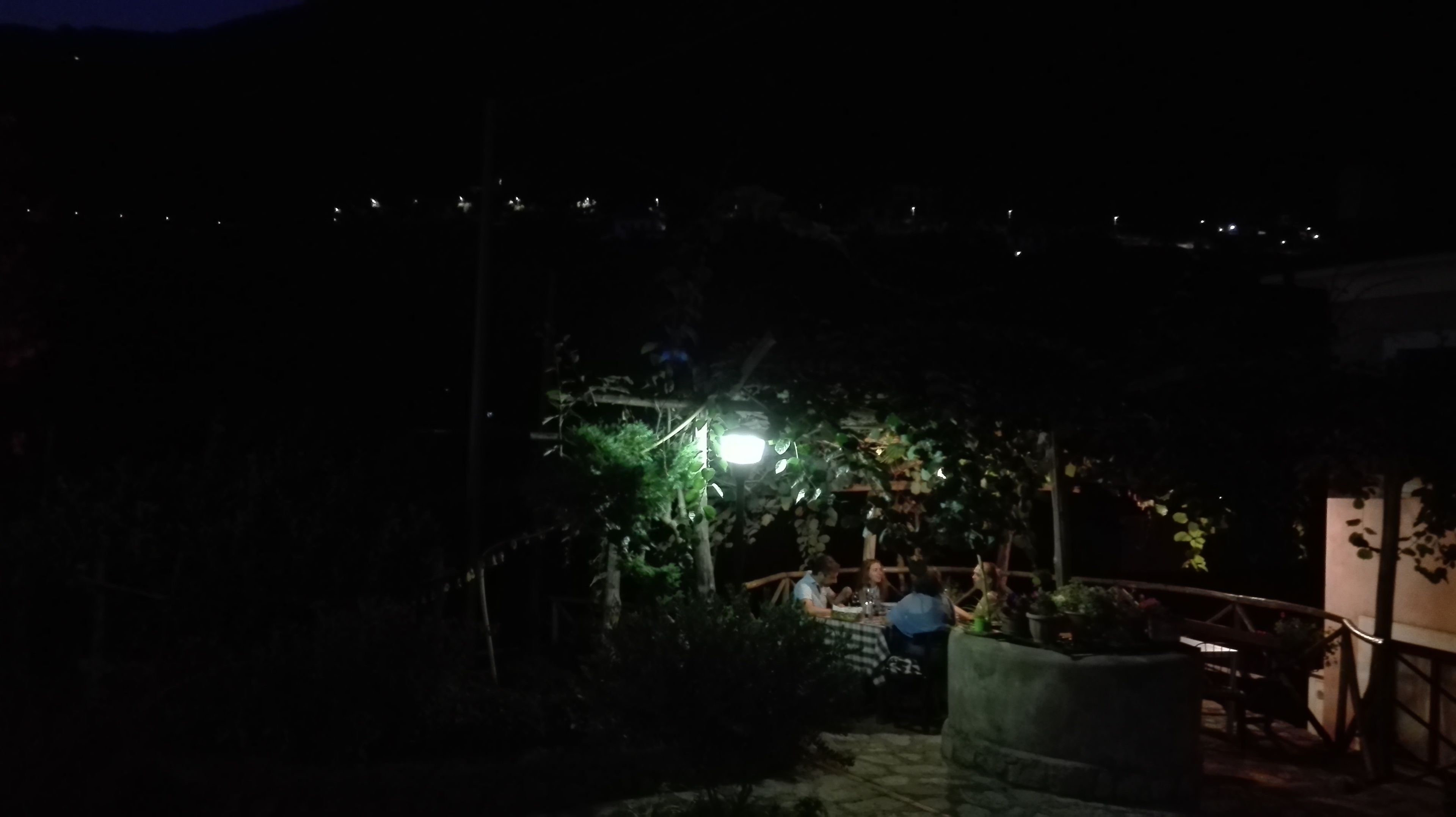 A dimly lit outdoor seating area with tables and chairs, surrounded by greenery and a small illuminated structure.