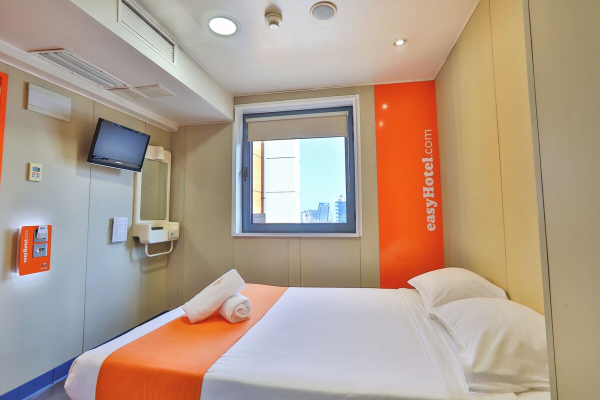 A hotel room with a bed, a television, a window, and a wall-mounted control panel.